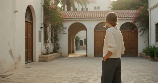 Woman in a minimalist neutral-toned outfit — a relaxed knit and tailored trousers, pausing at a sunlit Tel Aviv courtyard
