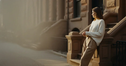 Woman in a minimalist neutral-toned outfit — a relaxed knit and tailored trousers, leaning against a Brooklyn brownstone s...
