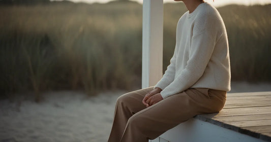 Woman in a minimalist neutral-toned outfit — a relaxed knit and tailored trousers, sitting on the edge of a Hamptons clapb...
