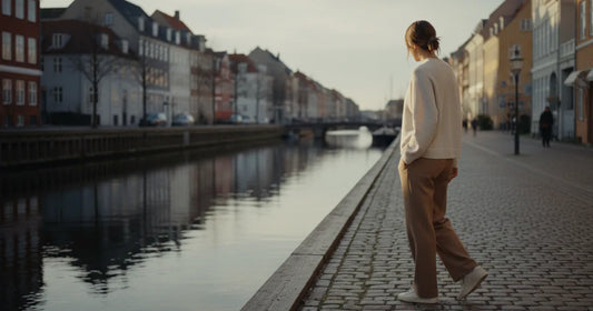 Woman in a minimalist neutral-toned outfit — a relaxed knit and tailored trousers, walking a quiet Copenhagen canal path a...