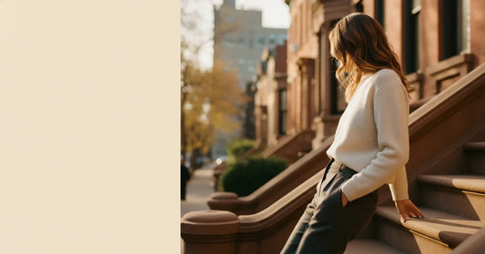 Woman in a minimalist neutral-toned outfit — a relaxed knit and tailored trousers, leaning against a Brooklyn brownstone s...