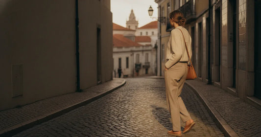 Woman in a minimalist neutral-toned outfit — a relaxed knit and tailored trousers, walking a Lisbon cobblestone alley at g...