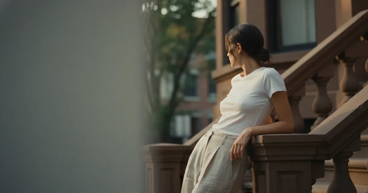 Woman in a perfectly-fitted white t-shirt and wide trousers, leaning against a Brooklyn brownstone stoop in late-afternoon...