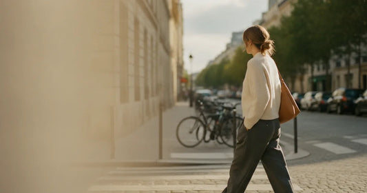 Woman in a minimalist neutral-toned outfit — a relaxed knit and tailored trousers, crossing a sunlit Paris side street in...