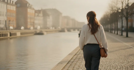 Woman in a minimalist neutral-toned outfit — a relaxed knit and tailored trousers, walking a quiet Copenhagen canal path a...