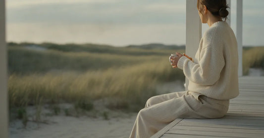 Woman in a minimalist neutral-toned outfit — a relaxed knit and tailored trousers, sitting on the edge of a Hamptons clapb...