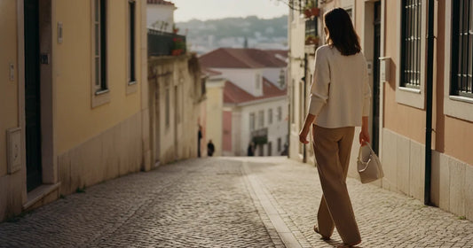 Woman in a minimalist neutral-toned outfit — a relaxed knit and tailored trousers, walking a Lisbon cobblestone alley at g...