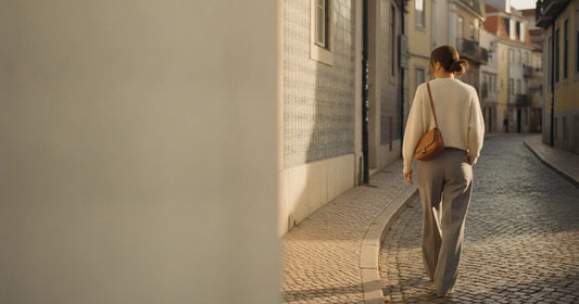 Woman in a minimalist neutral-toned outfit — a relaxed knit and tailored trousers, walking a Lisbon cobblestone alley at g...