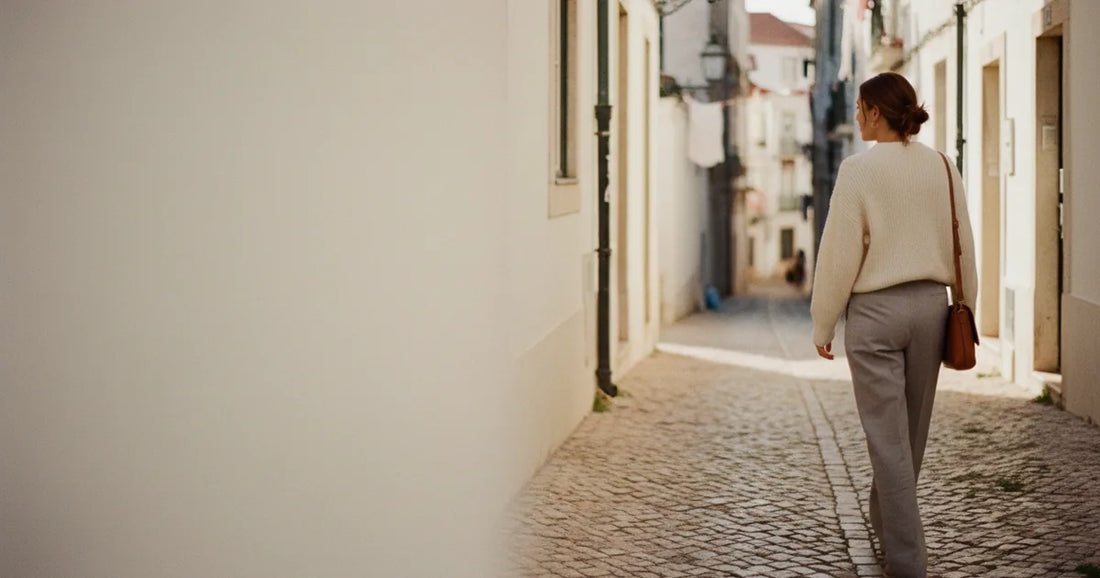Woman in a minimalist neutral-toned outfit — a relaxed knit and tailored trousers, walking a Lisbon cobblestone alley at g...