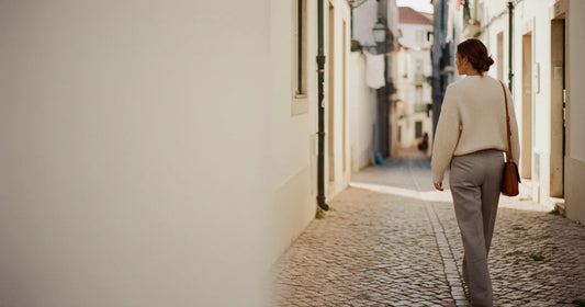Woman in a minimalist neutral-toned outfit — a relaxed knit and tailored trousers, walking a Lisbon cobblestone alley at g...