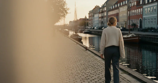 Woman in a minimalist neutral-toned outfit — a relaxed knit and tailored trousers, walking a quiet Copenhagen canal path a...