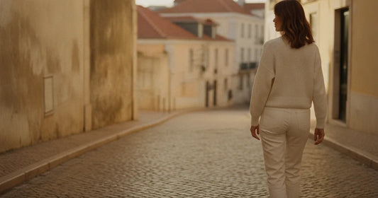 Woman in straight-leg ecru denim and a white knit, walking a Lisbon cobblestone alley at golden hour