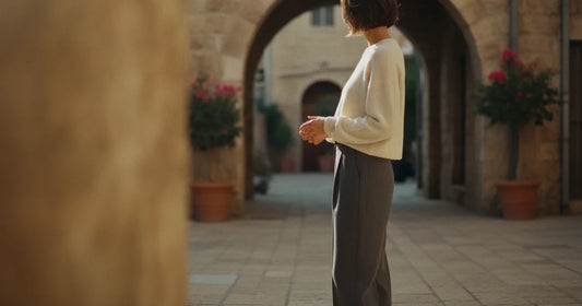Woman in a minimalist neutral-toned outfit — a relaxed knit and tailored trousers, pausing at a sunlit Tel Aviv courtyard