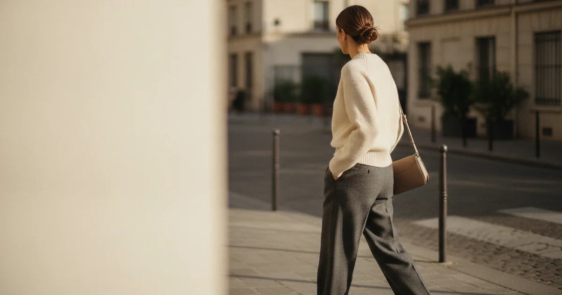 Woman in a minimalist neutral-toned outfit — a relaxed knit and tailored trousers, crossing a sunlit Paris side street in...