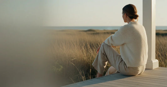 Woman in a minimalist neutral-toned outfit — a relaxed knit and tailored trousers, sitting on the edge of a Hamptons clapb...