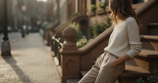 Woman in a minimalist neutral-toned outfit — a relaxed knit and tailored trousers, leaning against a Brooklyn brownstone s...
