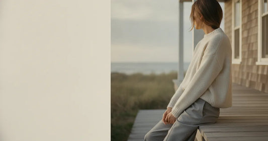 Woman in a minimalist neutral-toned outfit — a relaxed knit and tailored trousers, sitting on the edge of a Hamptons clapb...