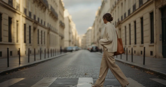 Woman in a minimalist neutral-toned outfit — a relaxed knit and tailored trousers, crossing a sunlit Paris side street in...
