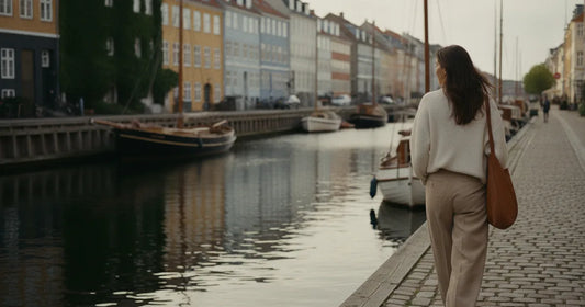 Woman in a minimalist neutral-toned outfit — a relaxed knit and tailored trousers, walking a quiet Copenhagen canal path a...
