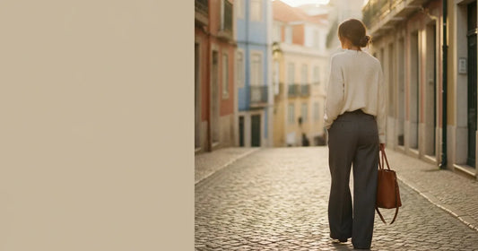Woman in a minimalist neutral-toned outfit — a relaxed knit and tailored trousers, walking a Lisbon cobblestone alley at g...