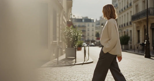 Woman in a minimalist neutral-toned outfit — a relaxed knit and tailored trousers, crossing a sunlit Paris side street in...
