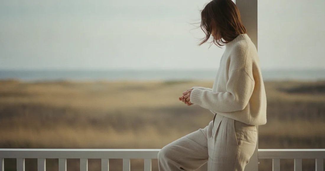 Woman in a minimalist neutral-toned outfit — a relaxed knit and tailored trousers, sitting on the edge of a Hamptons clapb...