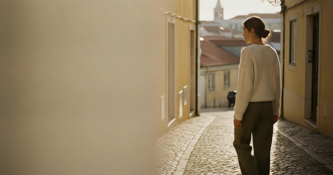 Woman in a minimalist neutral-toned outfit — a relaxed knit and tailored trousers, walking a Lisbon cobblestone alley at g...