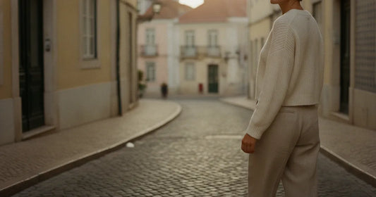 Woman in a minimalist neutral-toned outfit — a relaxed knit and tailored trousers, walking a Lisbon cobblestone alley at g...